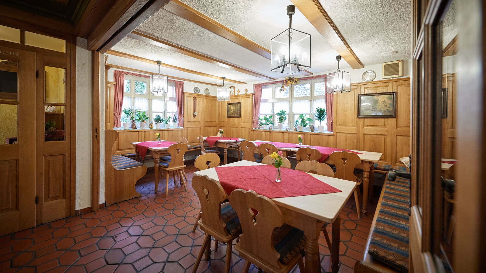Kleinere Gaststube mit Holzbänken, roten Tischläufern und Sprossenfenstern